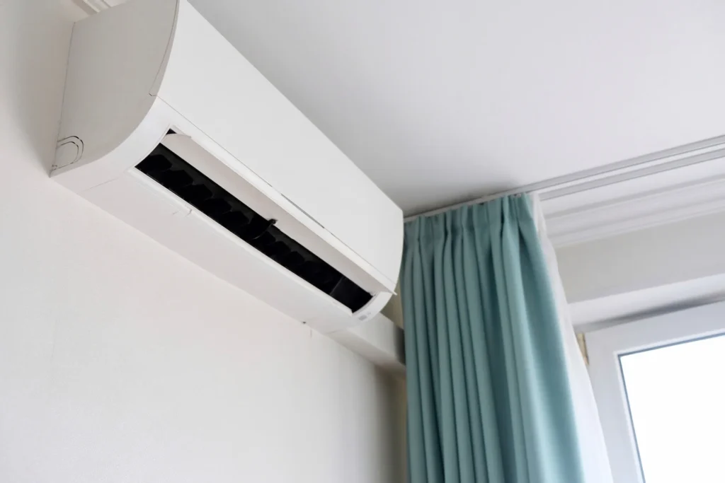 Modern air conditioner on white wall indoors. Hand with remote control directed on air conditioner.